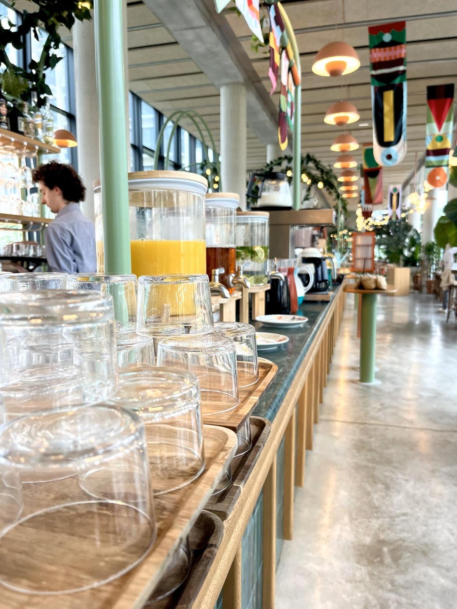 Auberge de jeunesse The People - Paris Marais Extérieur photo The photo shows a vibrant café or restaurant interior. In the foreground, there are several clear glass jars stacked on trays. Behind them, there are beverage dispensers — one contains orange juice, while another holds a different drink. The environm